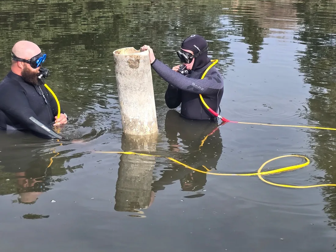 Two divers in wetsuits working on a submerged pipe in shallow water